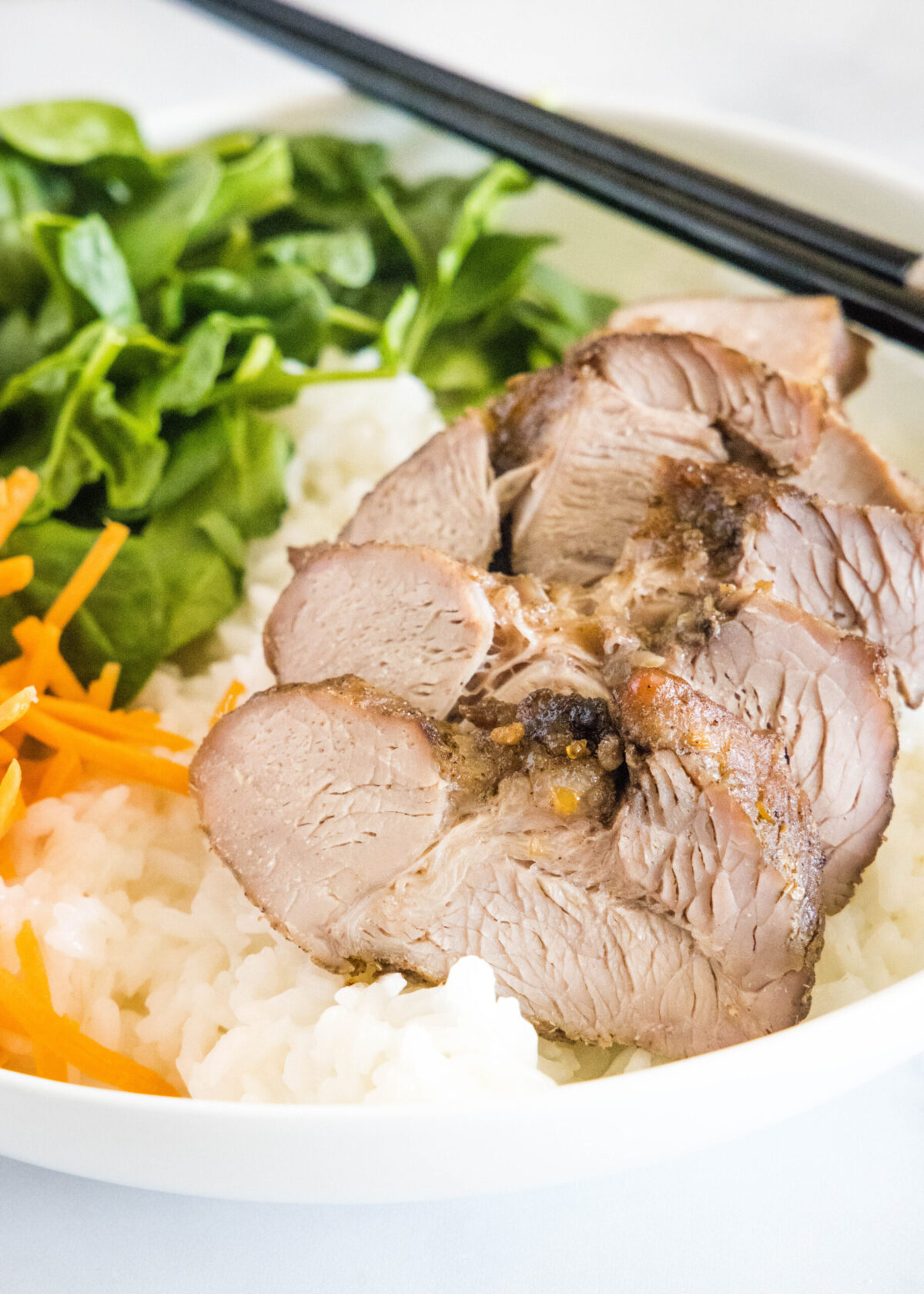 Close up of char siu pork in a bowl with rice, carrots, spinach, and chopsticks
