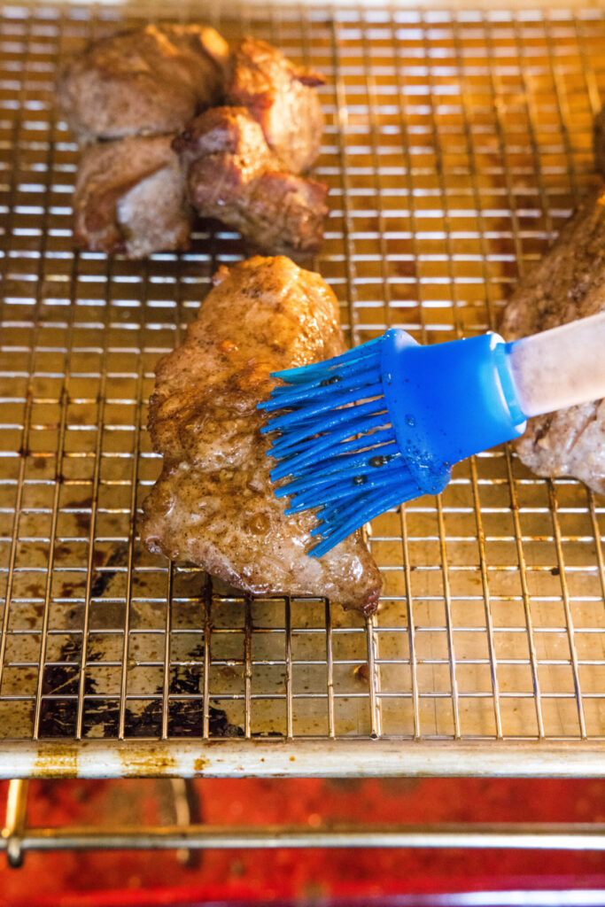 A kitchen brush painting sauce on top of pieces of pork on a wire rack