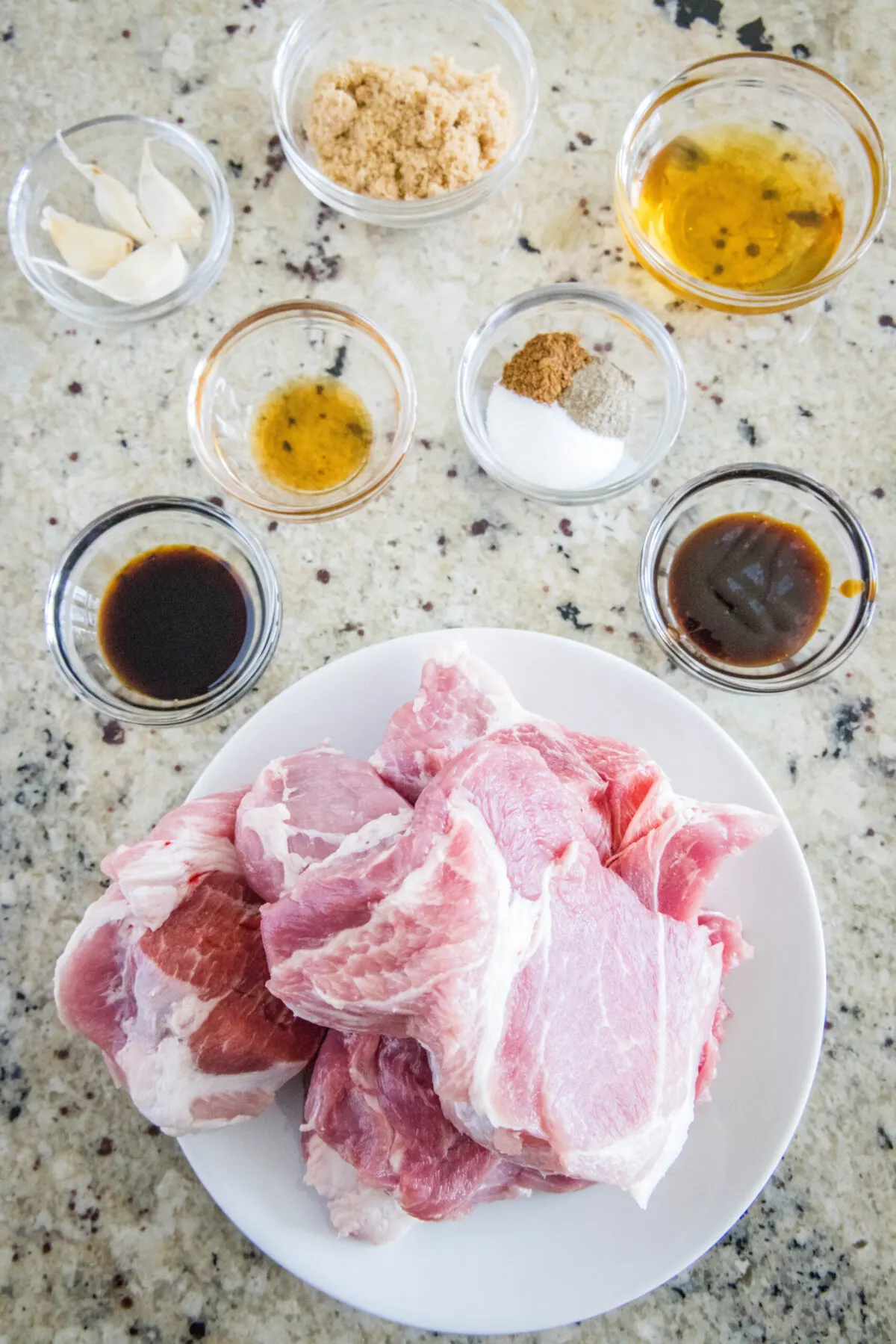 Overhead view of the ingredients needed for char siu pork: a plate of raw pork shoulder, a bowl of soy sauce, a bowl of hoisin, a bowl of garlic, a bowl of sesame oil, a bowl of brown sugar, a bowl of honey, and a bowl with salt, pepper, and five-spice.