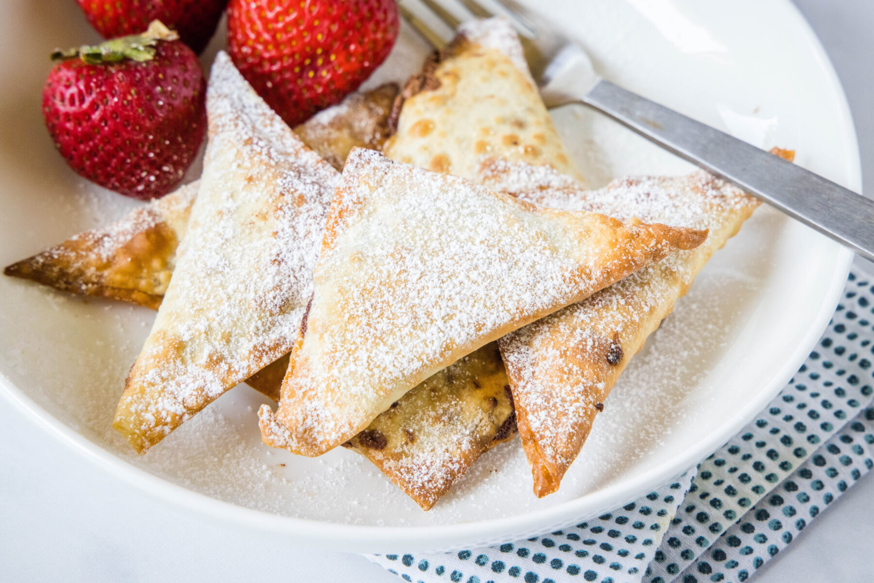 A plate of wontons covered in powdered sugar with strawberries and a fork