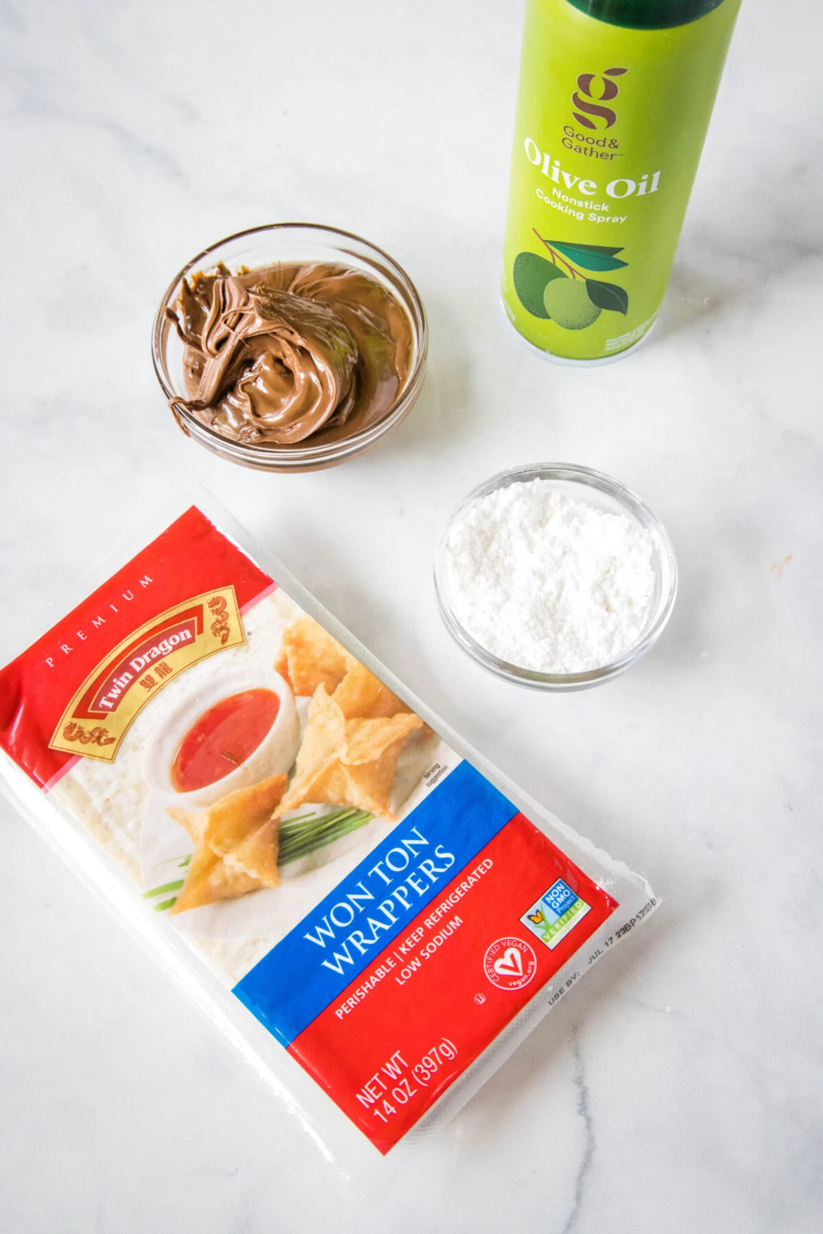 Overhead view of the ingredients needed for dessert wontons: a package of wonton wrappers, a bowl of Nutella, a bowl of powdered sugar, and a bottle of oil spray