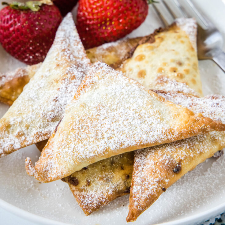 Close up of a pile of dessert wontons covered in powdered sugar next to strawberries and a fork
