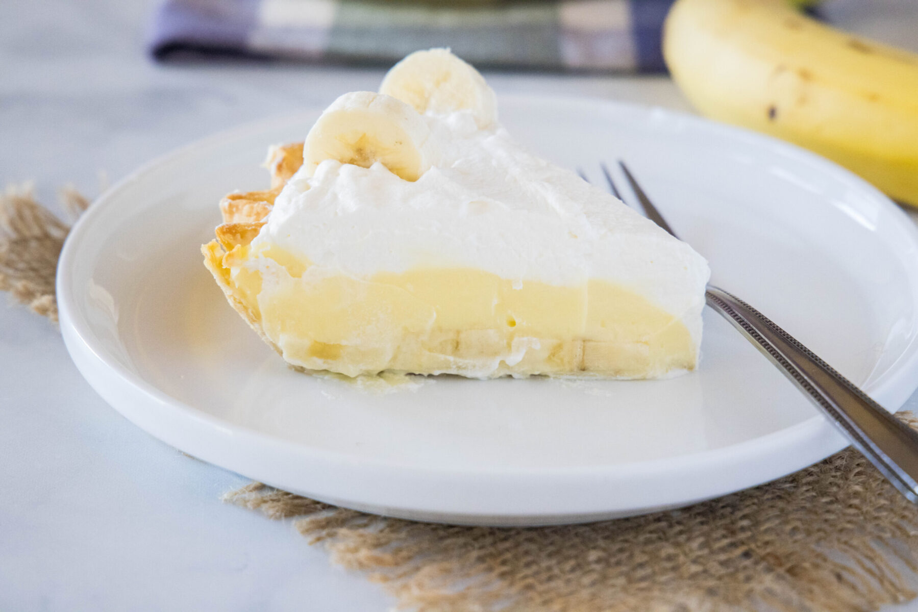 A slice of banana cream pie on a plate with a fork, with a banana in the background