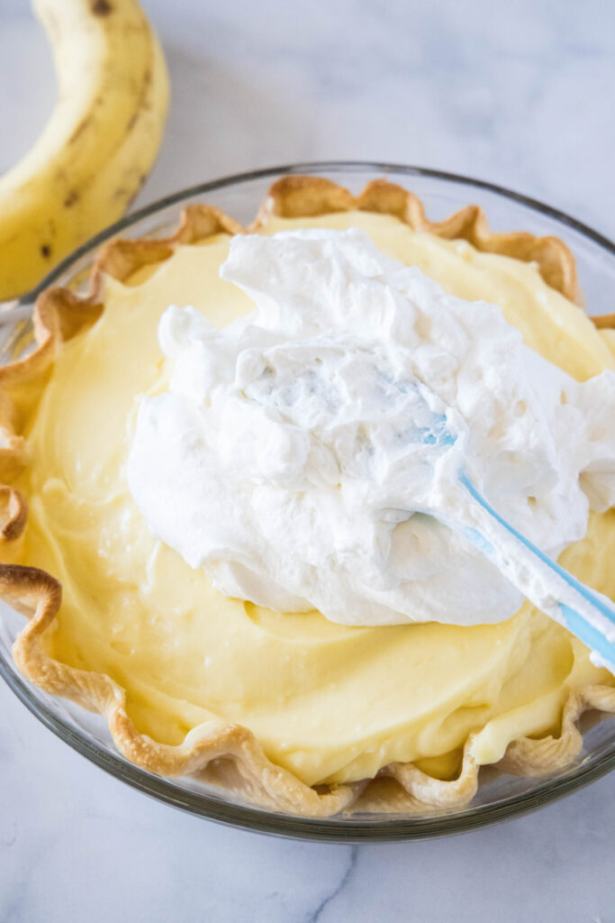 A spatula spreading whipped cream over the top of a banana cream pie
