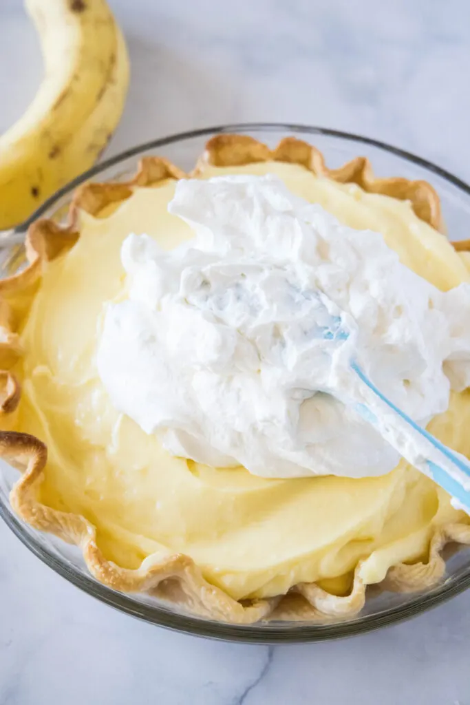 A spatula spreading whipped cream over the top of a banana cream pie
