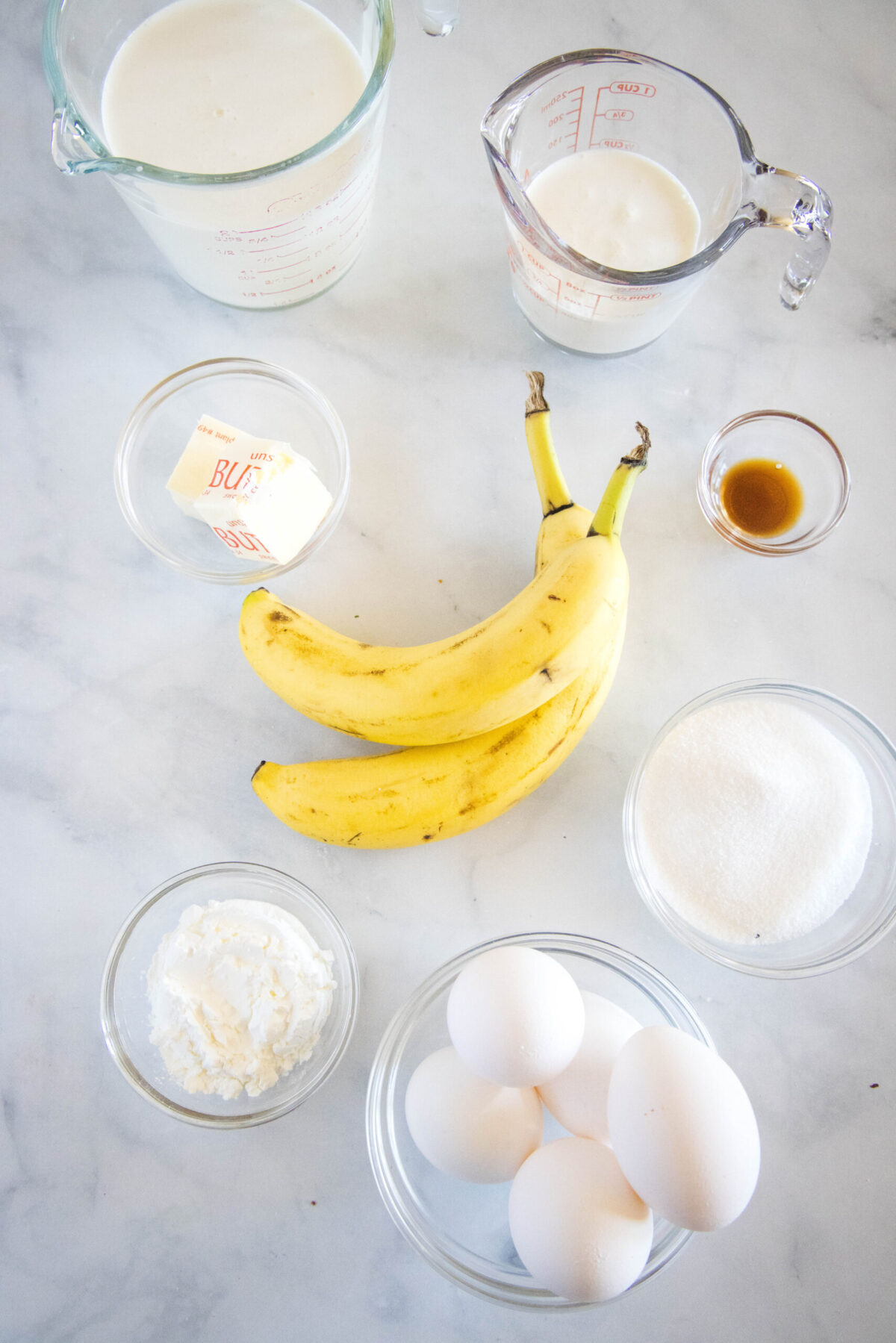 Overhead view of the ingredients needed for banana cream pie: a bowl of eggs, a bowl of sugar, a bowl of cornstarch, a bowl of butter, a bowl of milk, a bowl of cream, a bowl of vanilla, and two bananas