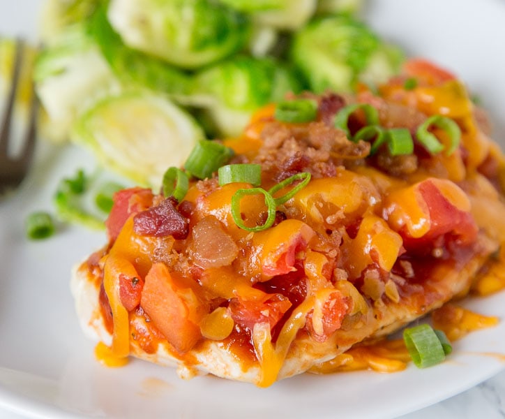 A close up of a plate of food with chicken topped with tomatoes, bacon and melted cheese
