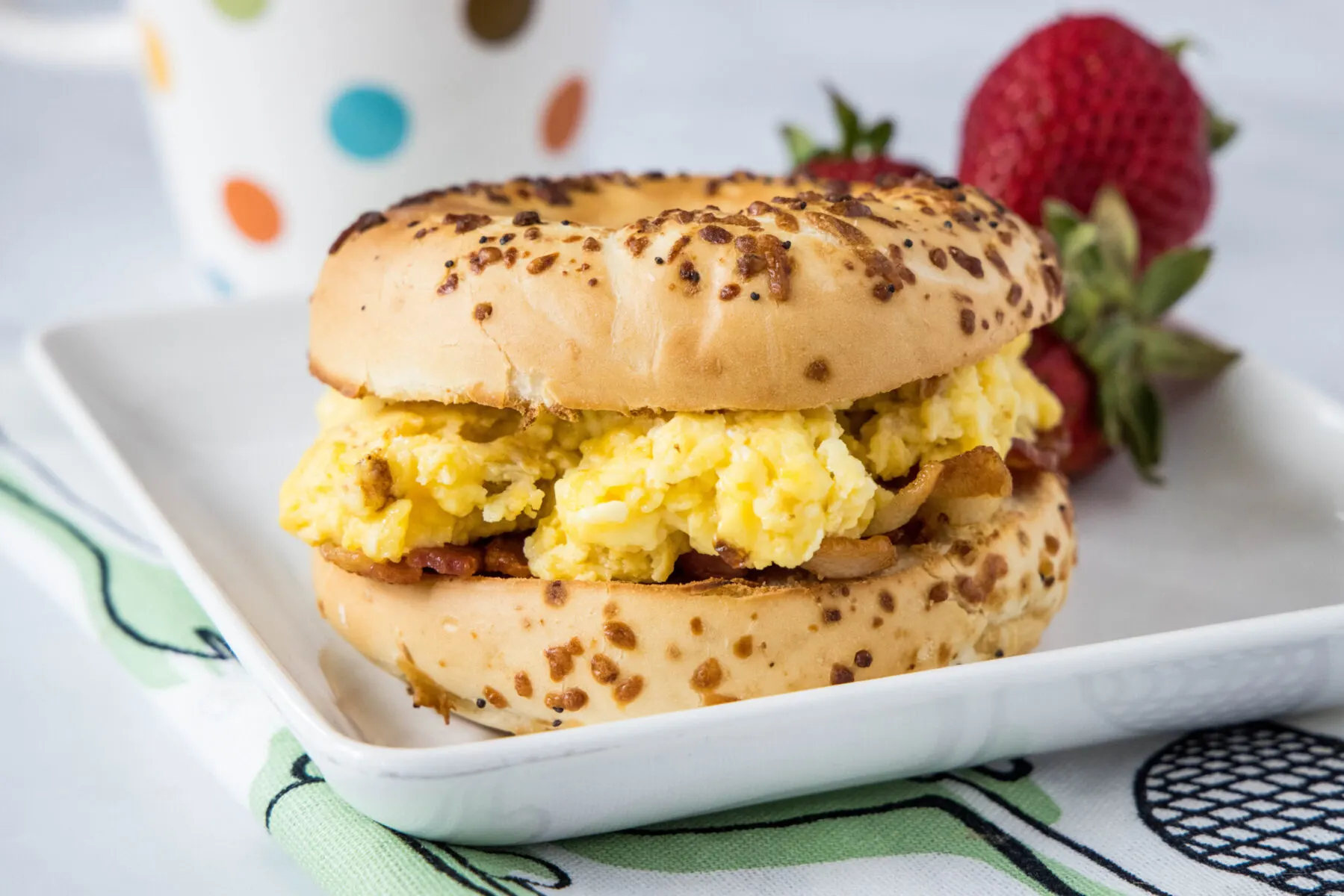 A bagel stuffed with eggs and bacon on a plate with strawberries and a mug in the background