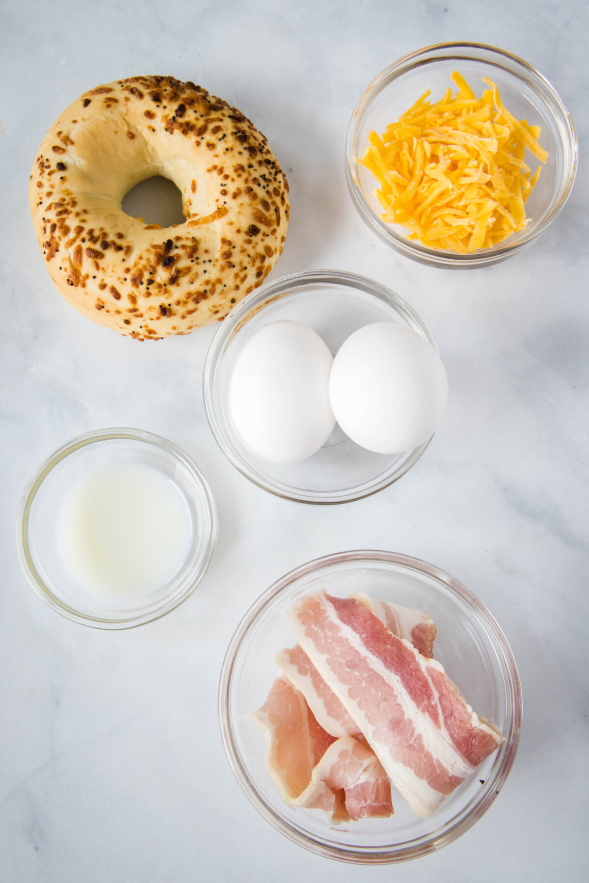 Overhead view of the ingredients needed for a bacon egg and cheese sandwich: a bagel, a bowl of bacon, a bowl of shredded cheese, a bowl of milk, and two eggs