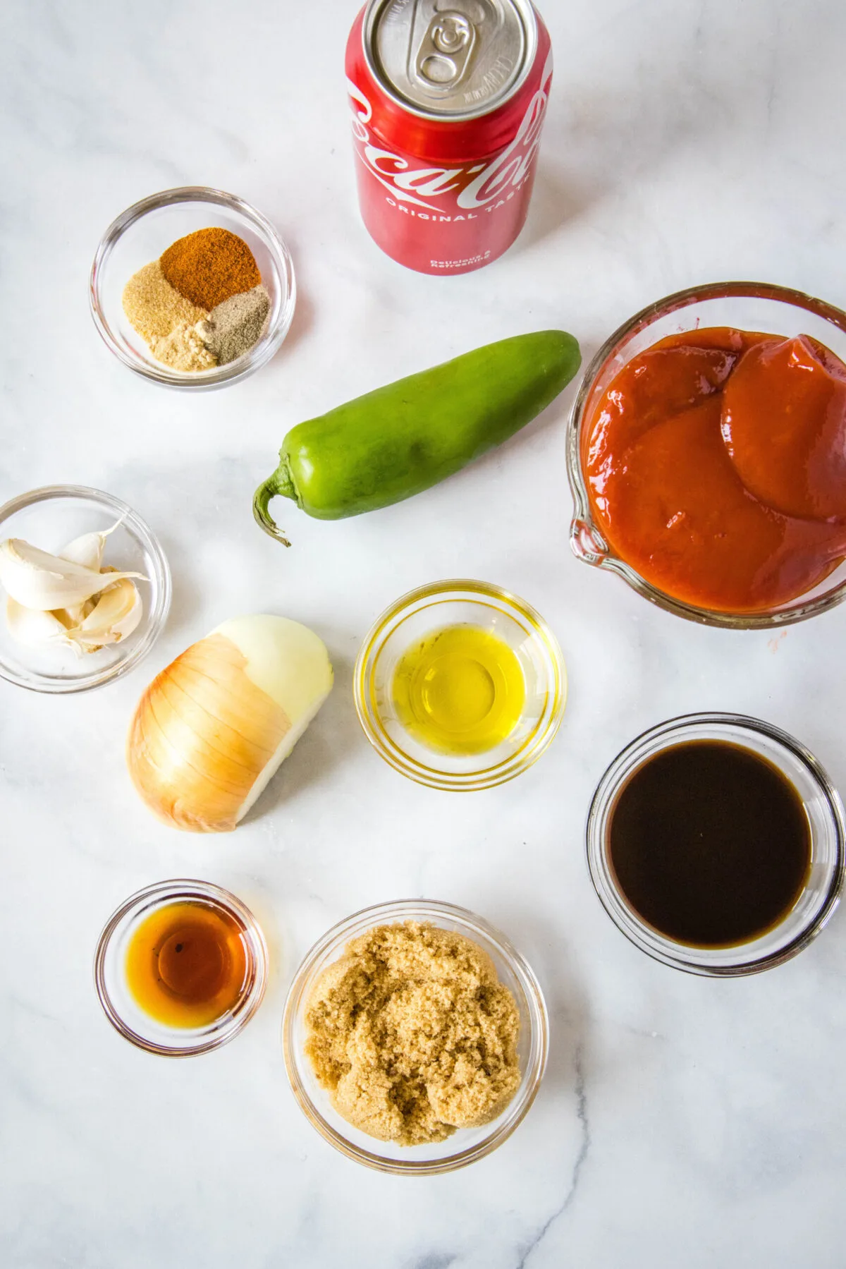 Overhead view of the ingredients needed for Coca-Cola BBQ sauce: a can of Coke, a bowl of ketchup, a jalapeño, a bowl of garlic, a bowl of oil, half an onion, a bowl of brown sugar, a bowl of Worcestershire sauce, a bowl of liquid smoke, and a bowl of onion powder, garlic powder, pepper, and paprika