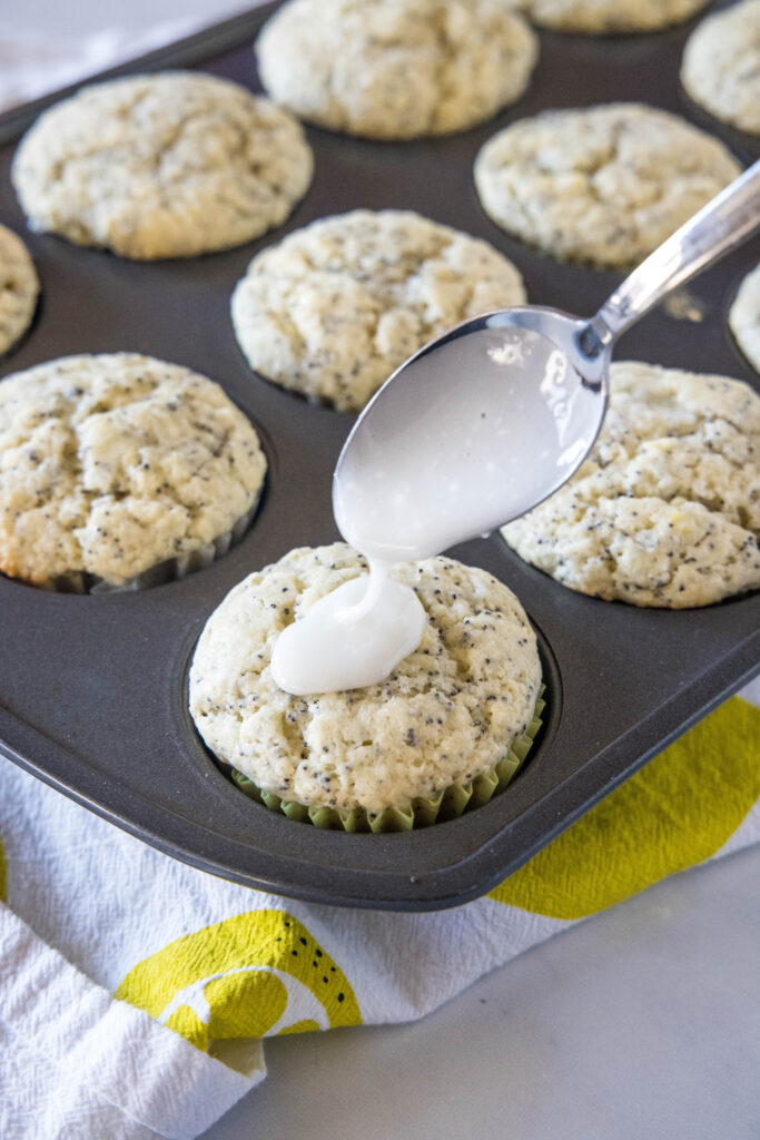A spoon drizzling glaze over a lemon poppy seed muffin in a muffin tin.