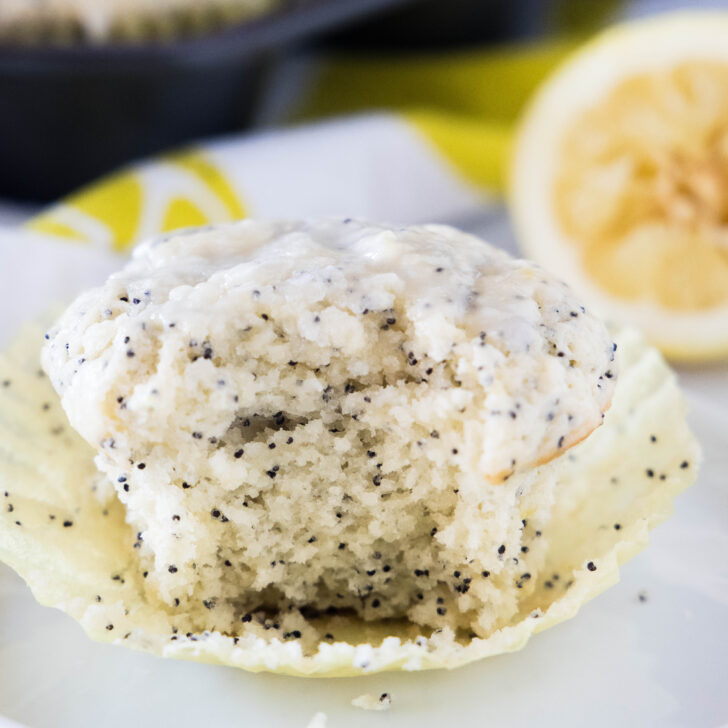 Close up of an unwrapped lemon poppy seed muffin on a white plate, with a bite missing.