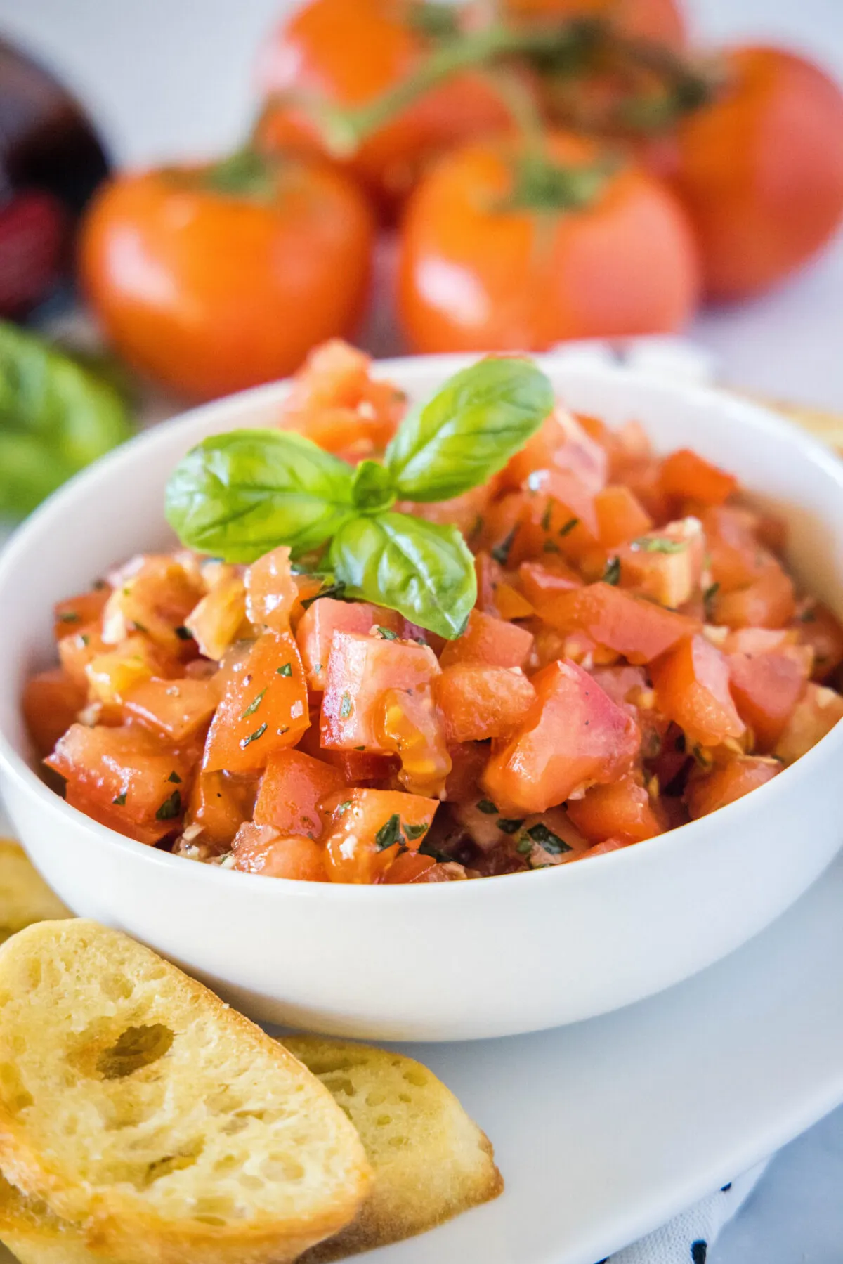 A bowl of bruschetta filling topped with basil, with bread slices in the foreground and tomatoes in the background