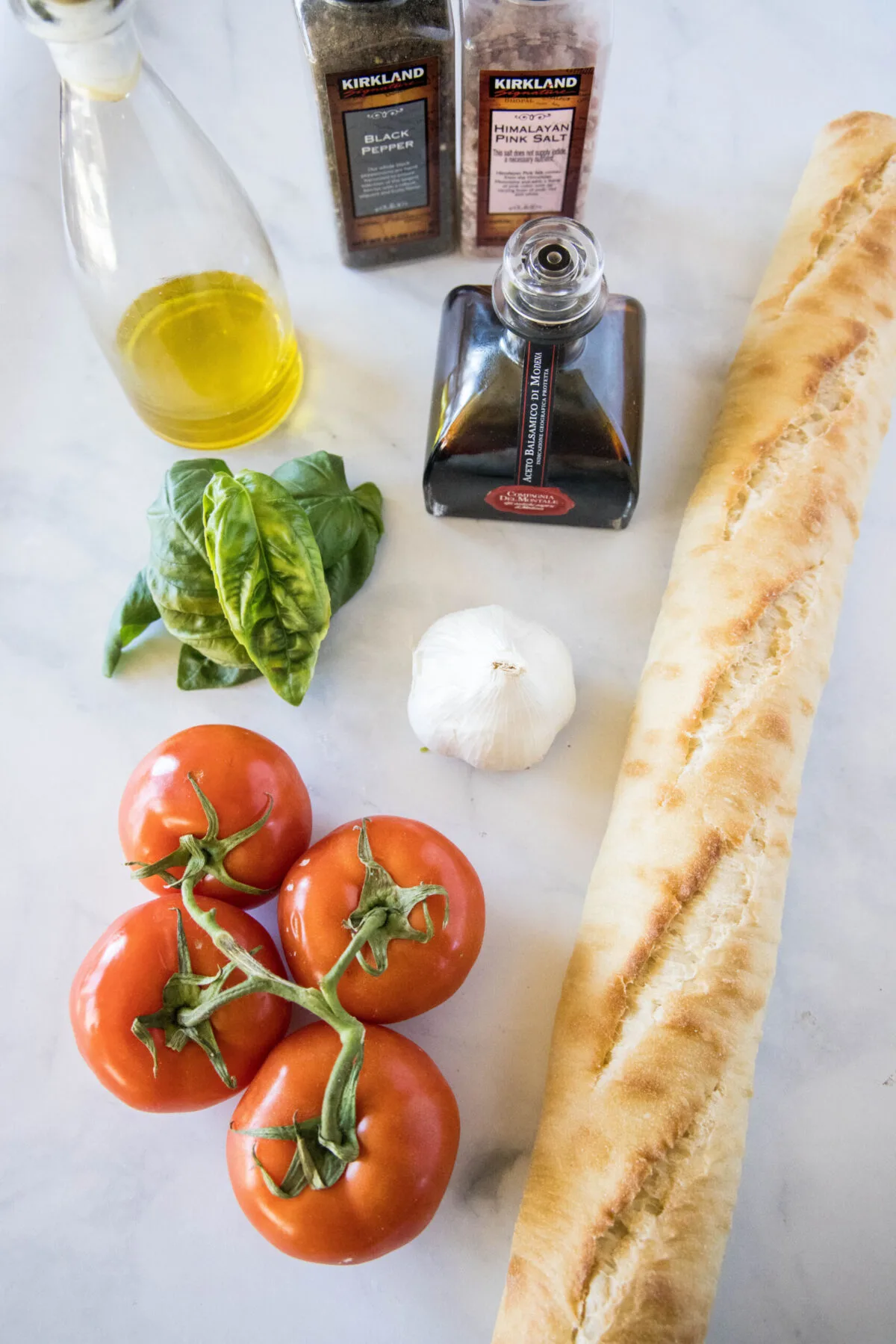 Overhead view of the ingredients needed for bruschetta: A baguette, four tomatoes on the vine, a head of garlic, a bunch of basil, a jar of balsamic vinegar, a bottle of olive oil, and salt and pepper