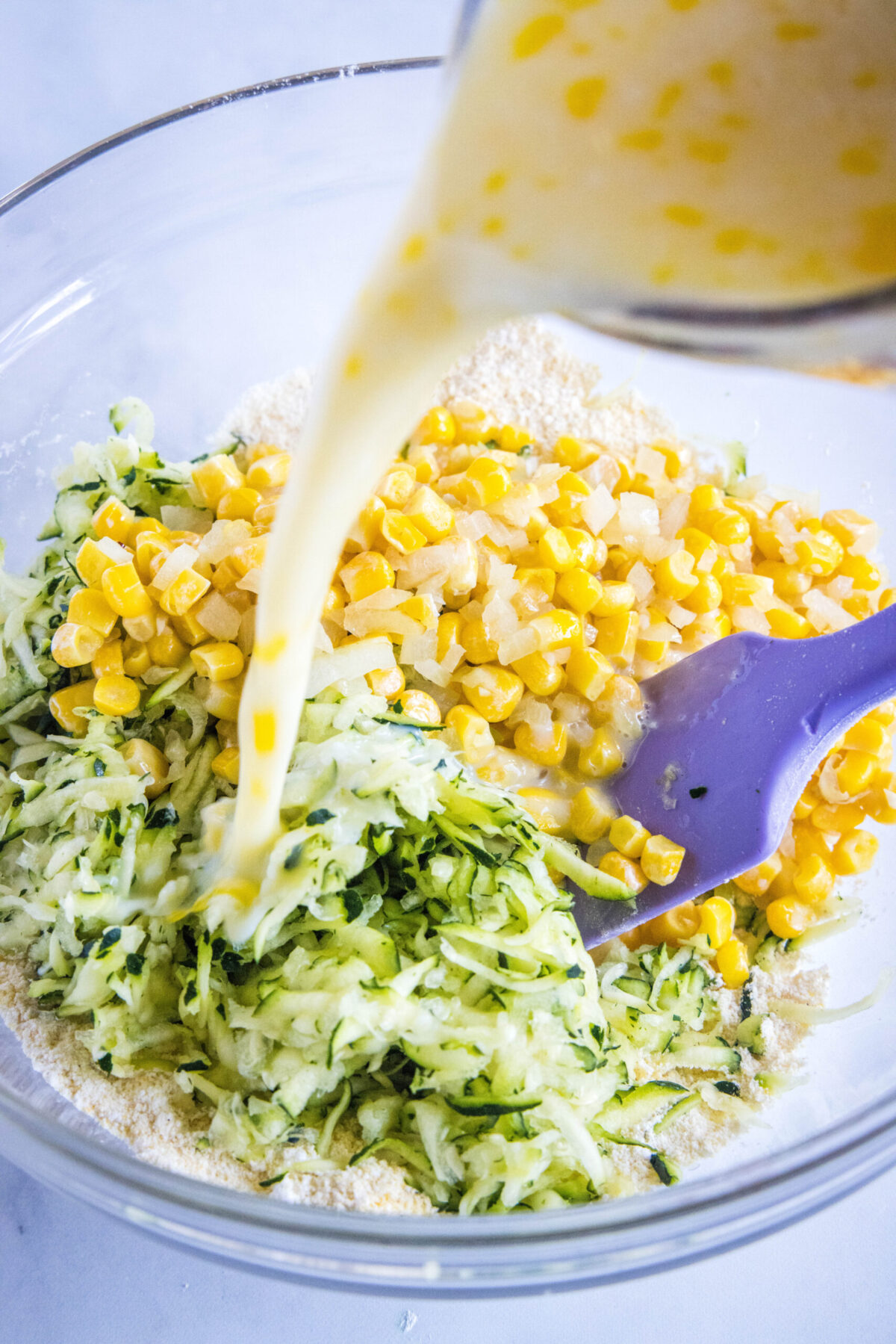 Buttermilk being poured into a mixing bowl with corn and shredded zucchini, with a spatula