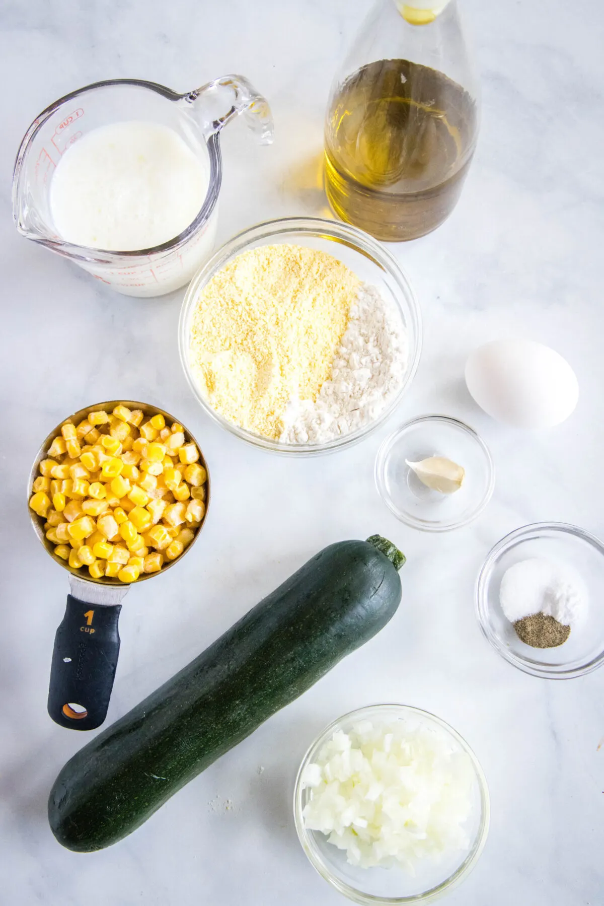 Overhead view of the ingredients needed for zucchini fritters with corn: A whole zucchini, a measuring cup of corn, a pyrex of buttermilk, a bowl of chopped onions, a bowl of cornmeal, an egg, a bowl of garlic, and a bowl of salt, pepper, and baking soda