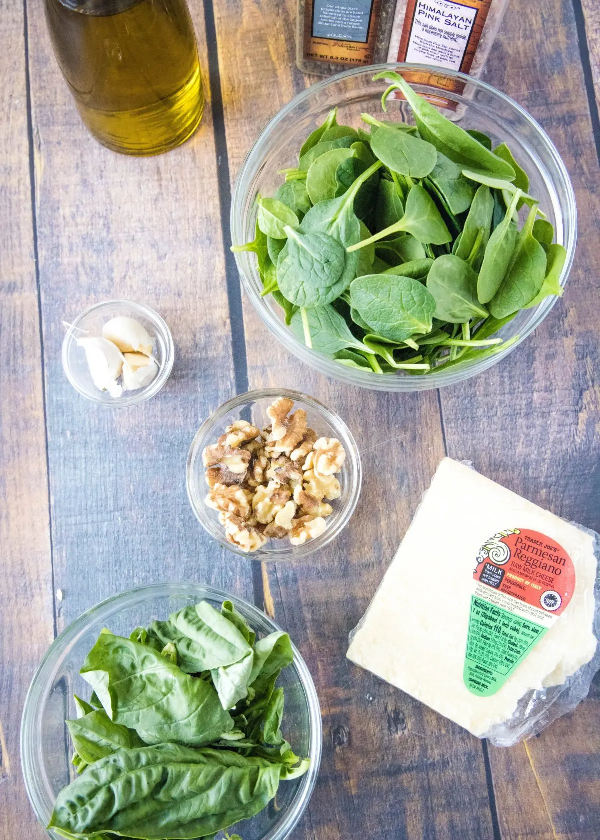 Overhead view of the ingredients needed for spinach pesto: a bowl of spinach, a bowl of basil, a bowl of walnuts, a bowl of garlic, a jar of olive oil, a block of parmesan cheese, and containers of salt and pepper