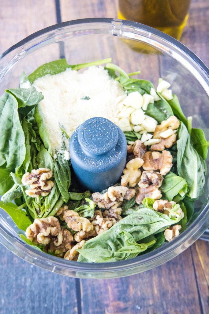 An overhead view of a food processor filled with garlic, parmesan cheese, spinach, and basil