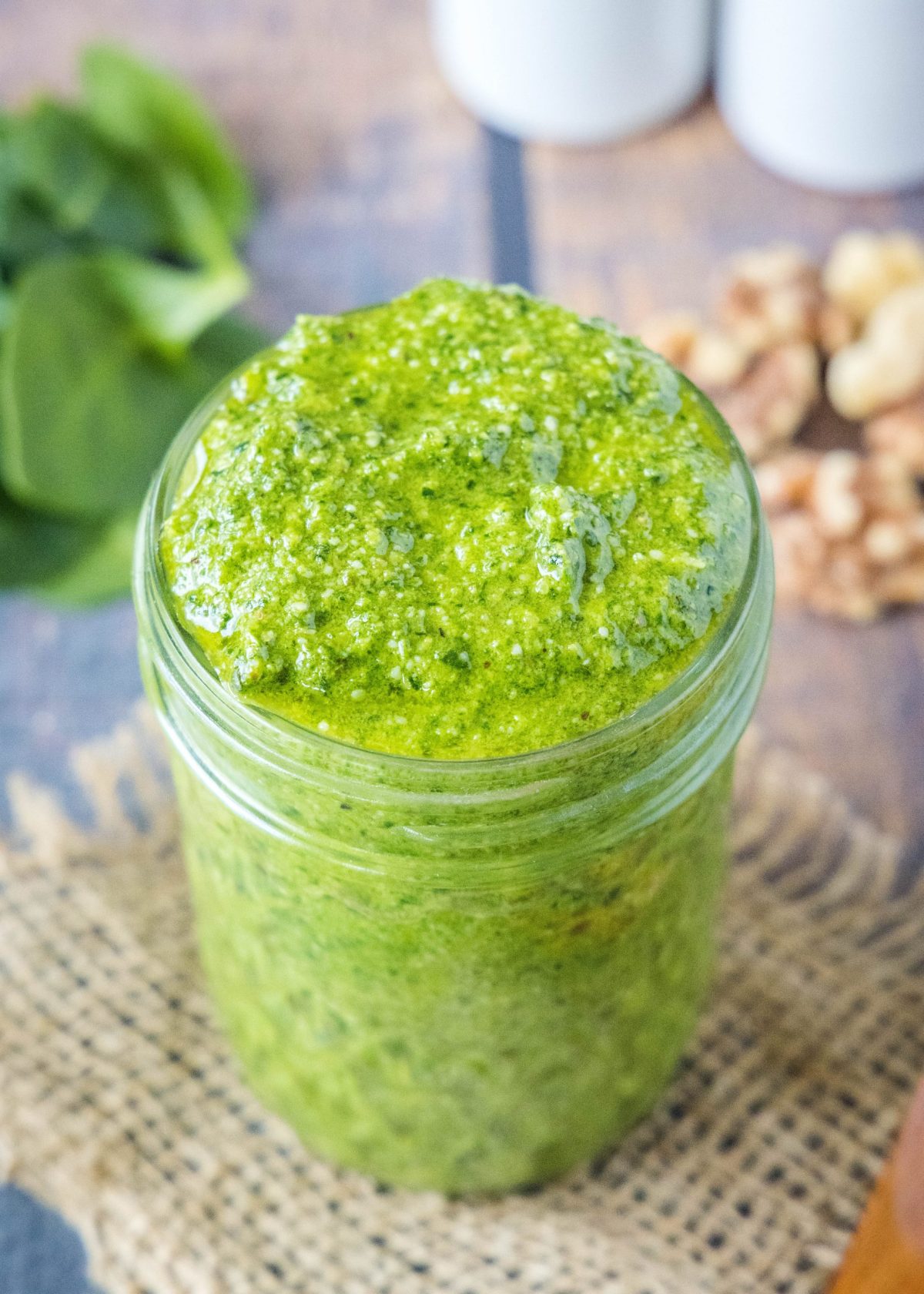 Close up of a jar of pesto with spinach and walnuts in the background