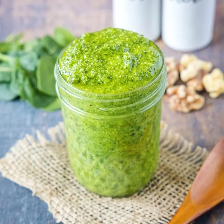 A jar of pesto with spinach and walnuts in the background