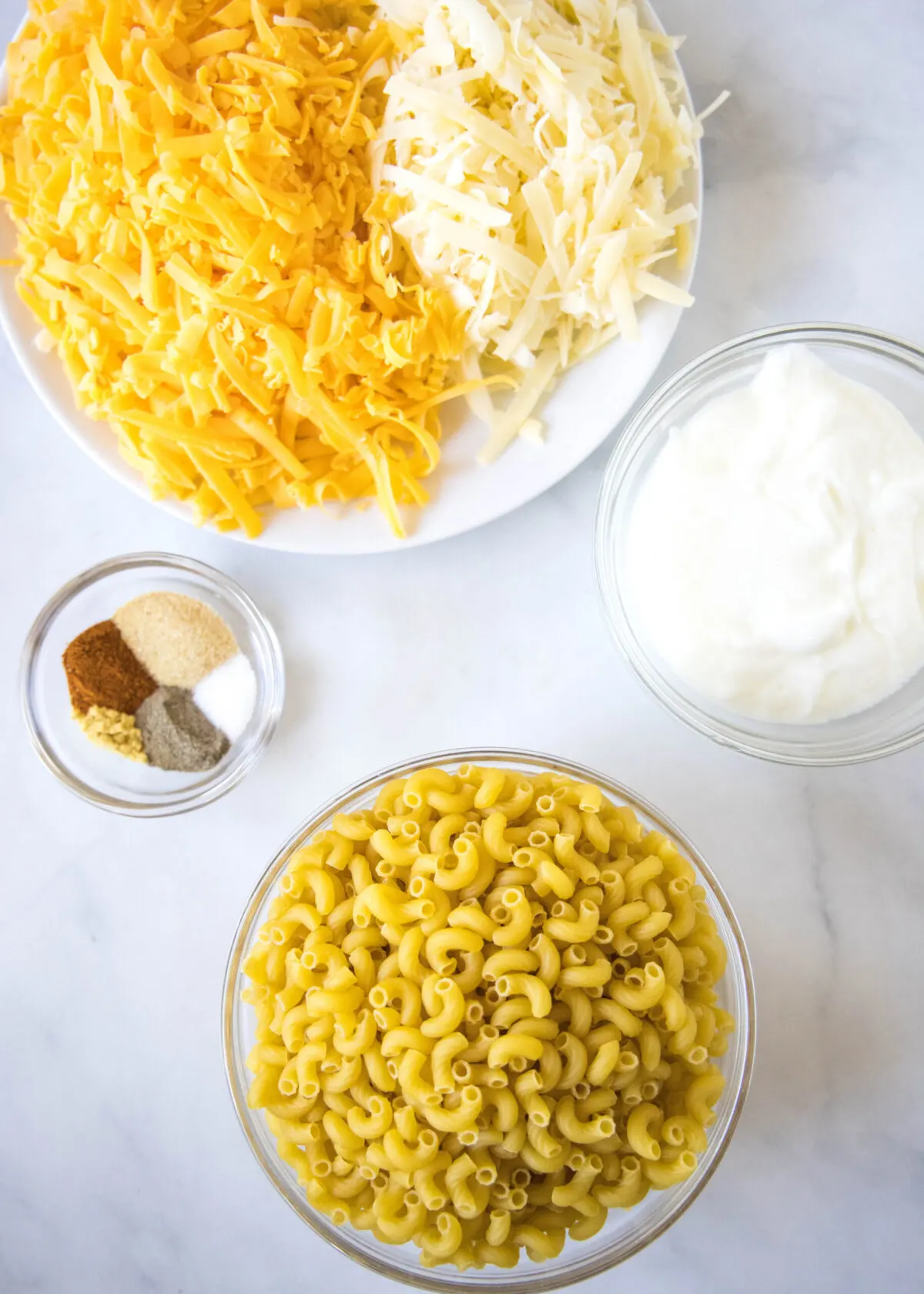 Overhead view of the ingredients needed for healthy mac and cheese: a bowl of elbow macaroni, a bowl of shredded gruyere and cheddar cheeses, a bowl of Greek yogurt, and a bowl of garlic powder, ground mustard, paprika, pepper, and salt