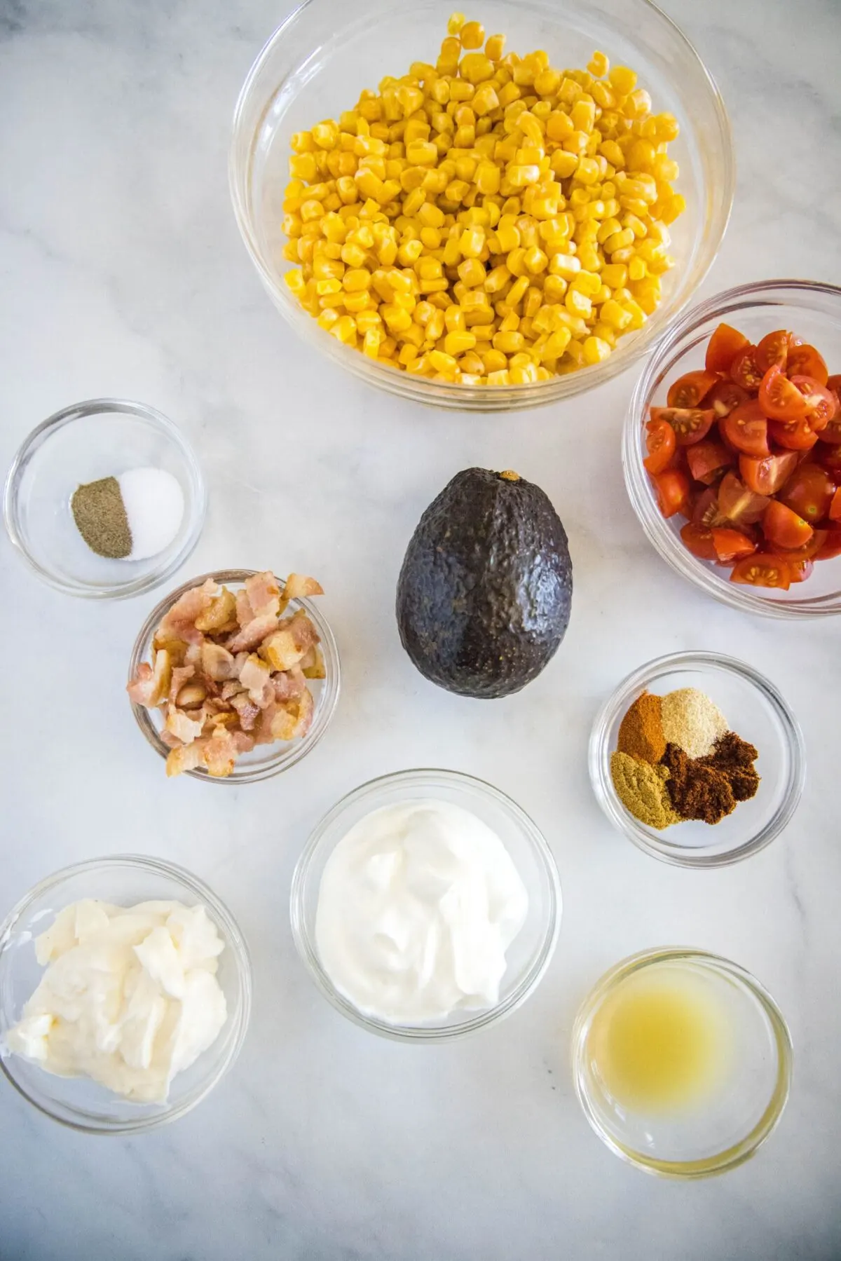 Overhead view of the ingredients needed for southwestern corn salad: a bowl of corn, a bowl of quartered cherry tomatoes, a bowl of cooked bacon crumbles, a bowl of salt and pepper, a bowl of greek yogurt, a bowl of mayo, a bowl of lime juice, a bowl of chili powder, cumin, garlic powder, and black pepper, and an avocado