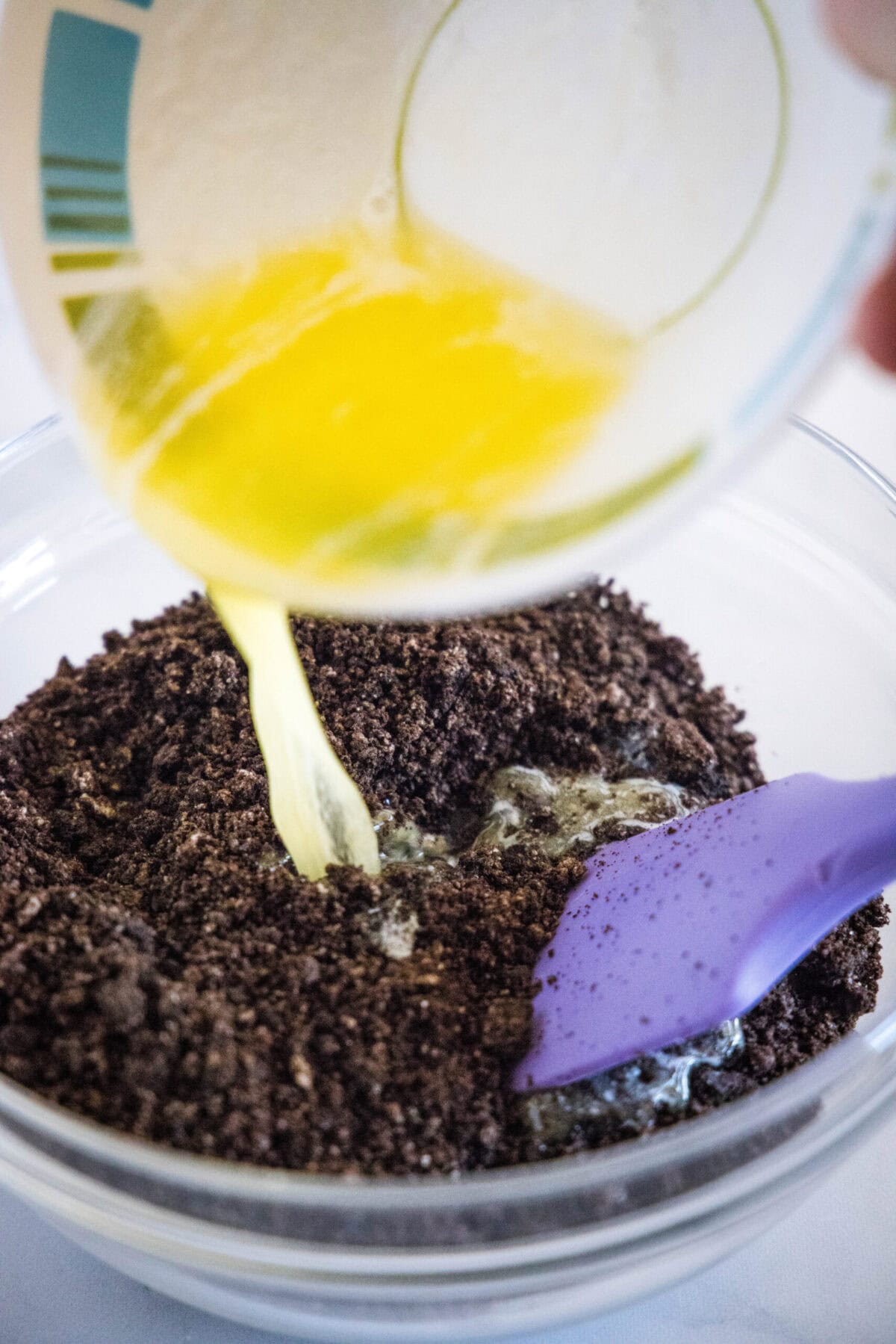 A pyrex pouring melted butter into a bowl of Oreo crumbs with a rubber spatula