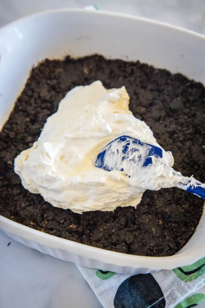 A rubber spatula spreading a cream cheese mixture over an Oreo crust in a baking dish
