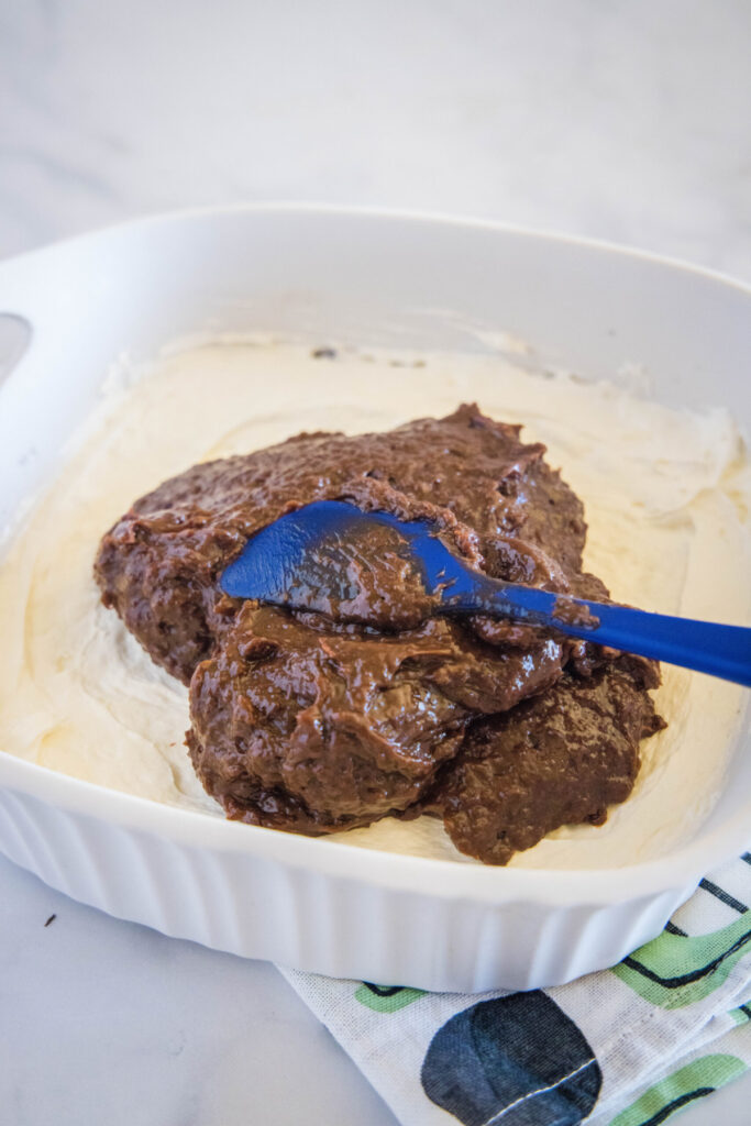 A rubber spatula spreading chocolate pudding over a cream cheese mixture in a baking dish