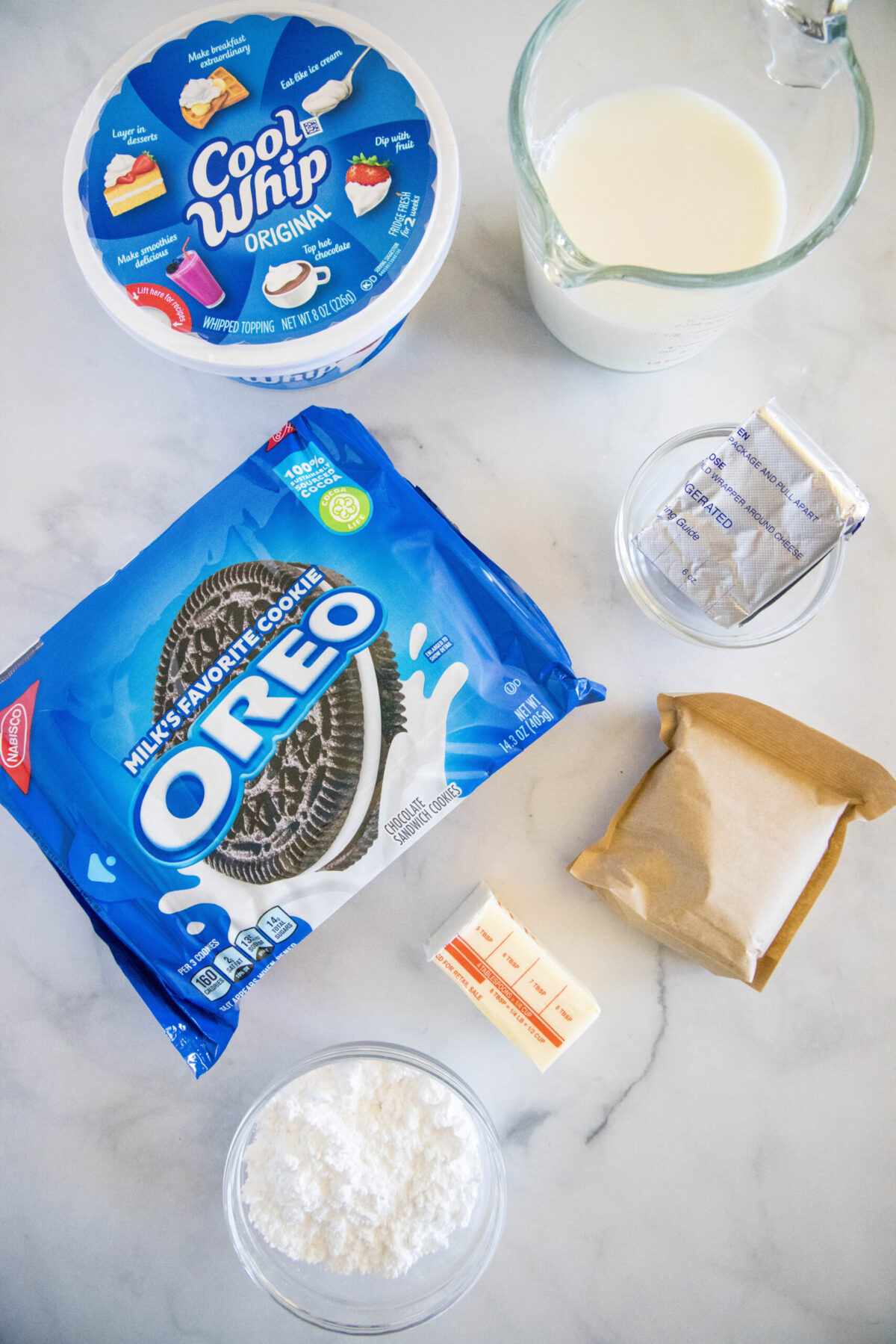 Overhead view of the ingredients needed for no bake Oreo cream squares: a pack of Oreos, a tub of Cool Whip, a pack of chocolate pudding mix, a pyrex of milk, a bowl of cream cheese, a bowl of powdered sugar, and half a stick of butter