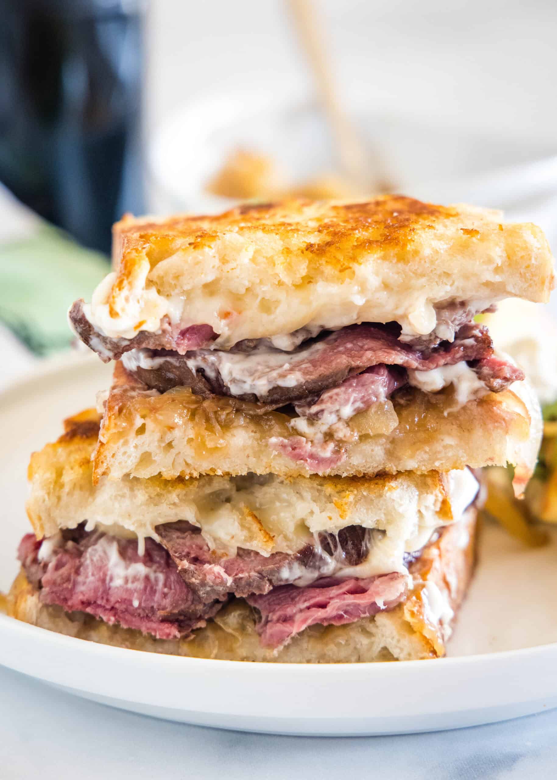Tuck into the best steak sandwich with juicy sliced beef, caramelized onions, garlic aioli, and melty cheese between two toasty bread slices! Two halves of a steak sandwich stacked on a white plate.