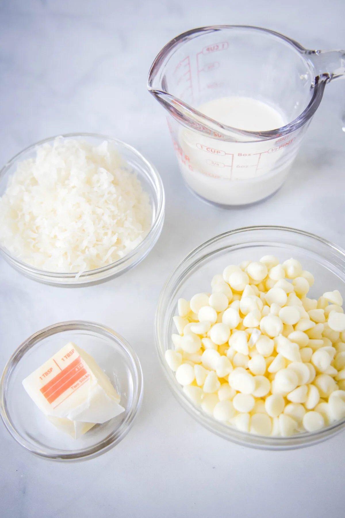 Overhead view of some of the ingredients needed for coconut balls: a bowl of white chocolate chips, a pyrex of heavy cream, a bowl of shredded coconut, and some butter