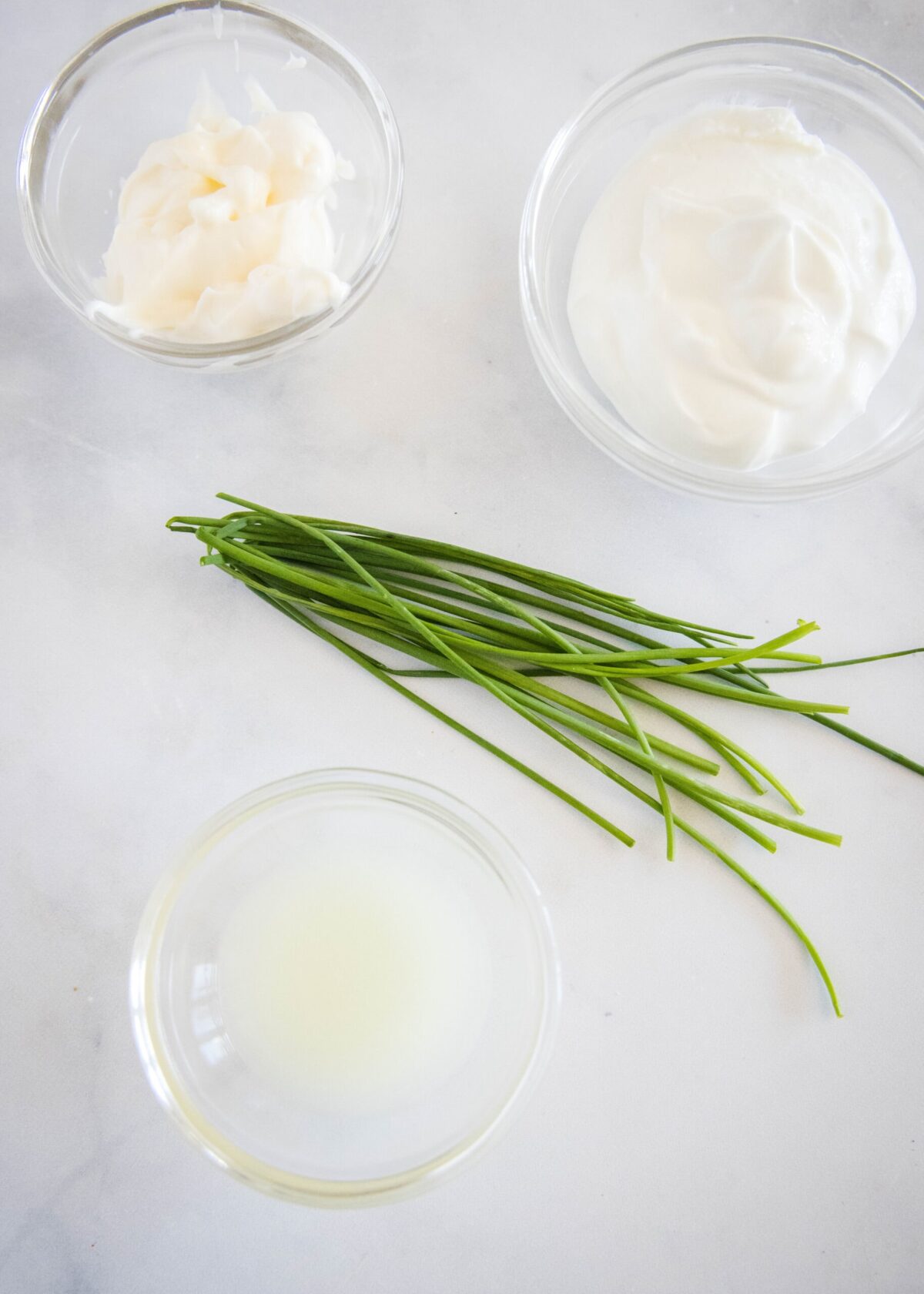 Overhead view of the ingredients needed for a dipping sauce: a handful of chives, a bowl of lemon juice, a bowl of mayo, and a bowl of Greek yogurt