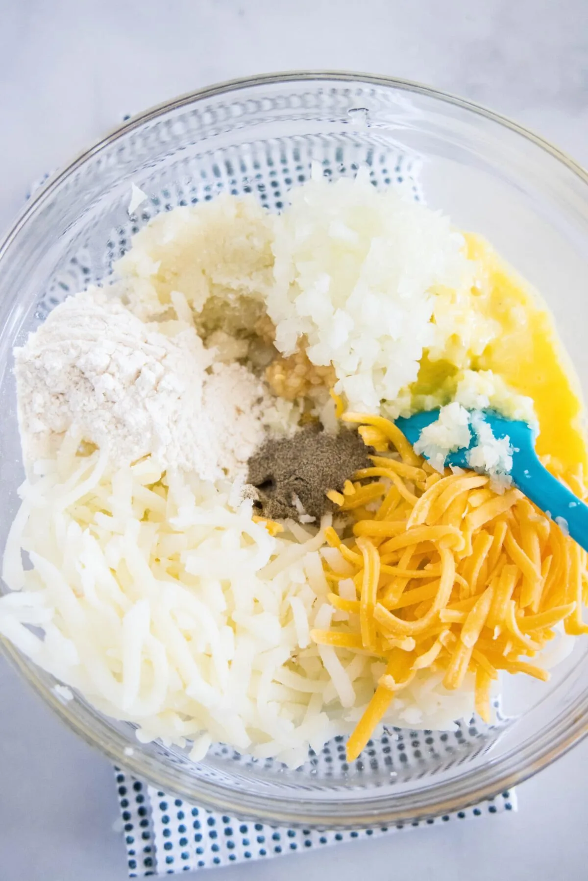 A mixing bowl with mashed cauliflower, shredded hash browns, grated cheese, onion, garlic, salt, pepper, an egg, and a rubber spatula