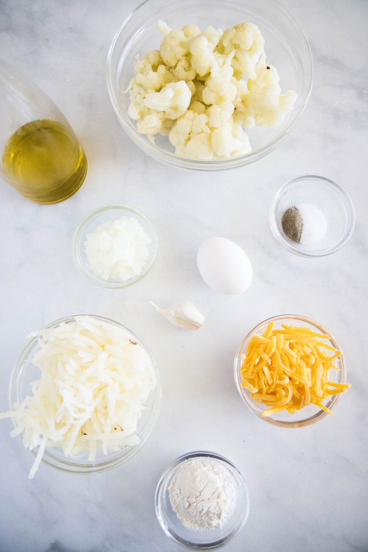 Overhead view of the ingredients needed to make cauliflower fritters: a bowl of cauliflower florets, a bowl of shredded hash browns, a bowl of grated cheddar cheese, a glass of olive oil, a bowl of salt and pepper, a bowl of chopped onion, a bowl of flour, a garlic clove, and an egg