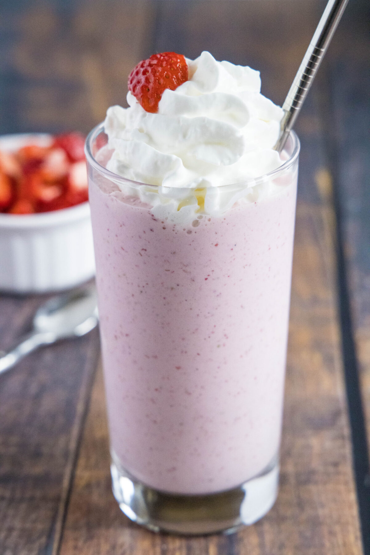 Close up of a glass of strawberry milkshake with whipped cream, a strawberry garnish, and a straw