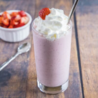 Wide angle view of a strawberry milkshake topped with whipped cream, next to a spoon and a bowl of strawberries.