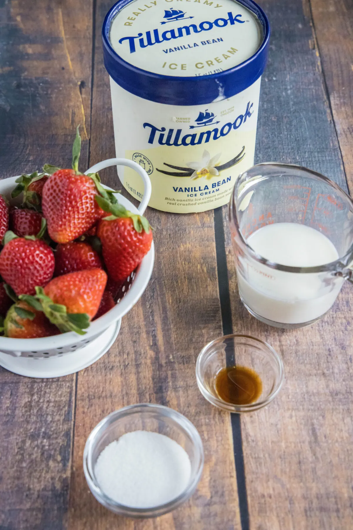 A counter with a bowl of fresh strawberries, a carton of Tillamook vanilla ice cream, a bowl of sugar, a bowl of vanilla, and a pyrex of milk