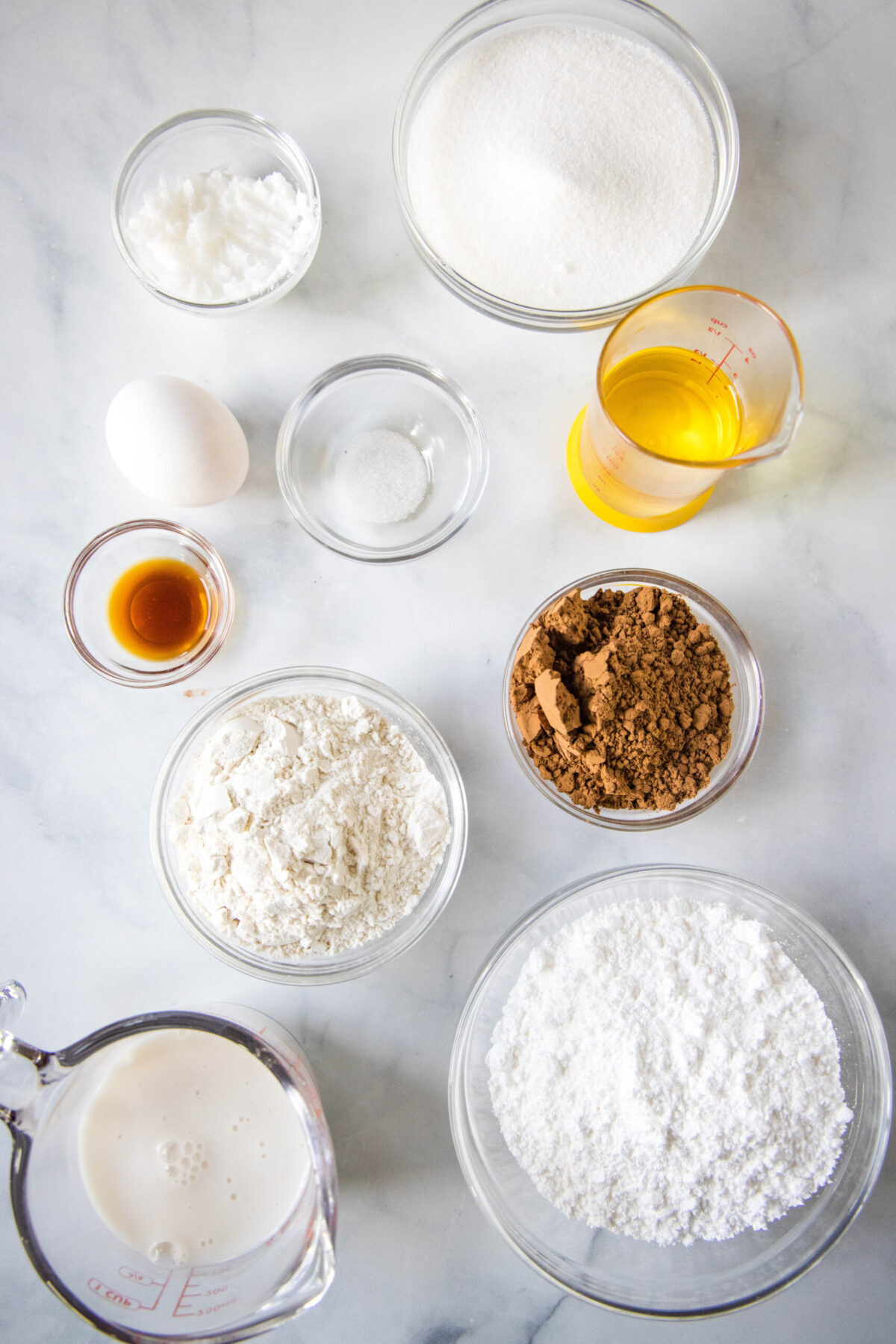 Overhead view of the ingredients needed for dairy free brownies: a pyrex of almond milk, a bowl of flour, a bowl of sugar, a bowl of cocoa powder, a glass of oil, a bowl of vanilla, a bowl of salt, an egg, and a bowl of coconut oil.