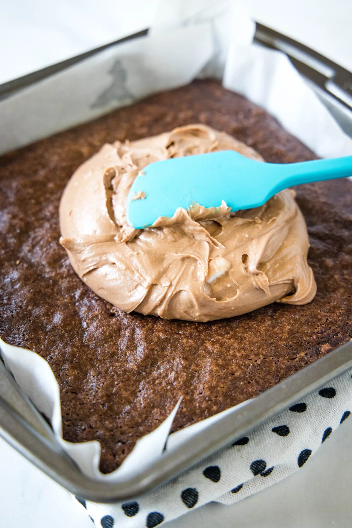 A rubber spatula spreading chocolate frosting over a pan of brownies.