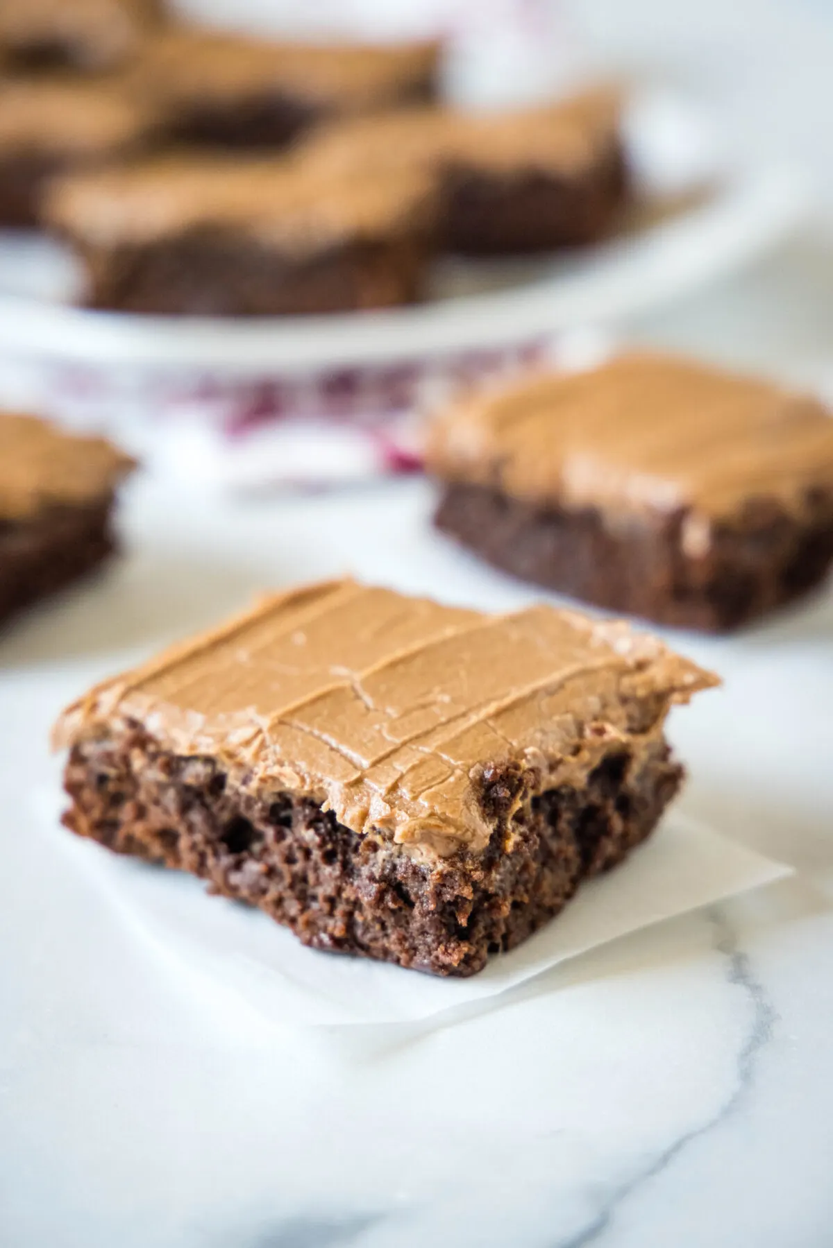 A brownie square with chocolate frosting, with a plate of brownies in the background.