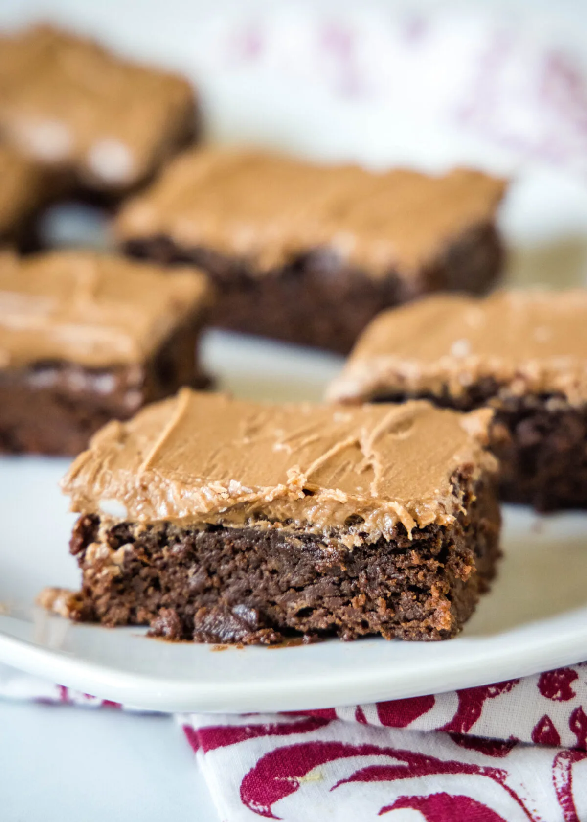 Close up of a brownie square with chocolate frosting, with more brownie squares in the background.