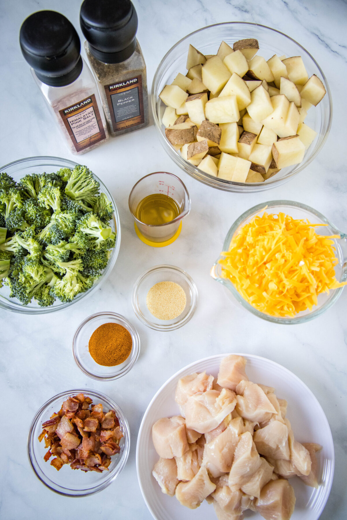 Overhead view of the ingredients needed for loaded chicken potato casserole: a bowl of diced potatoes, a bowl of broccoli, a bowl of cubed chicken breast, a bowl of bacon crumbles, a bowl of shredded cheese, a bowl of paprika, a bowl of garlic powder, a bowl of olive oil, and a salt and pepper shaker.