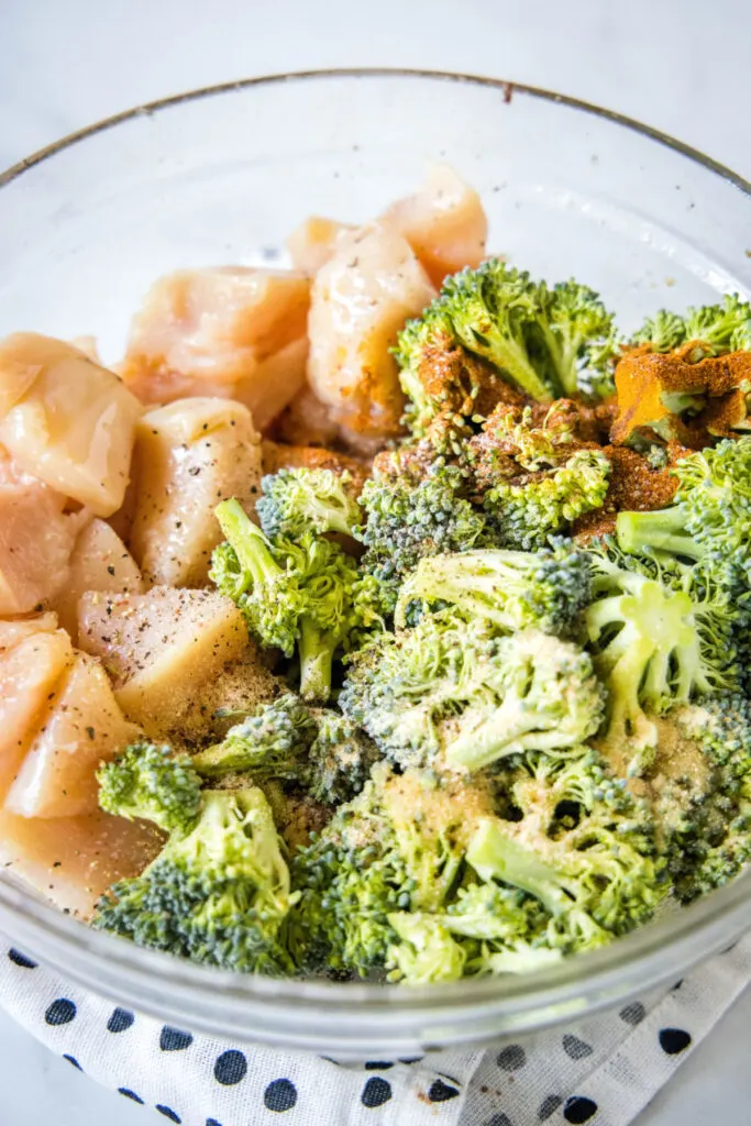 A mixing bowl with raw broccoli and chicken, topped with oil and spices.