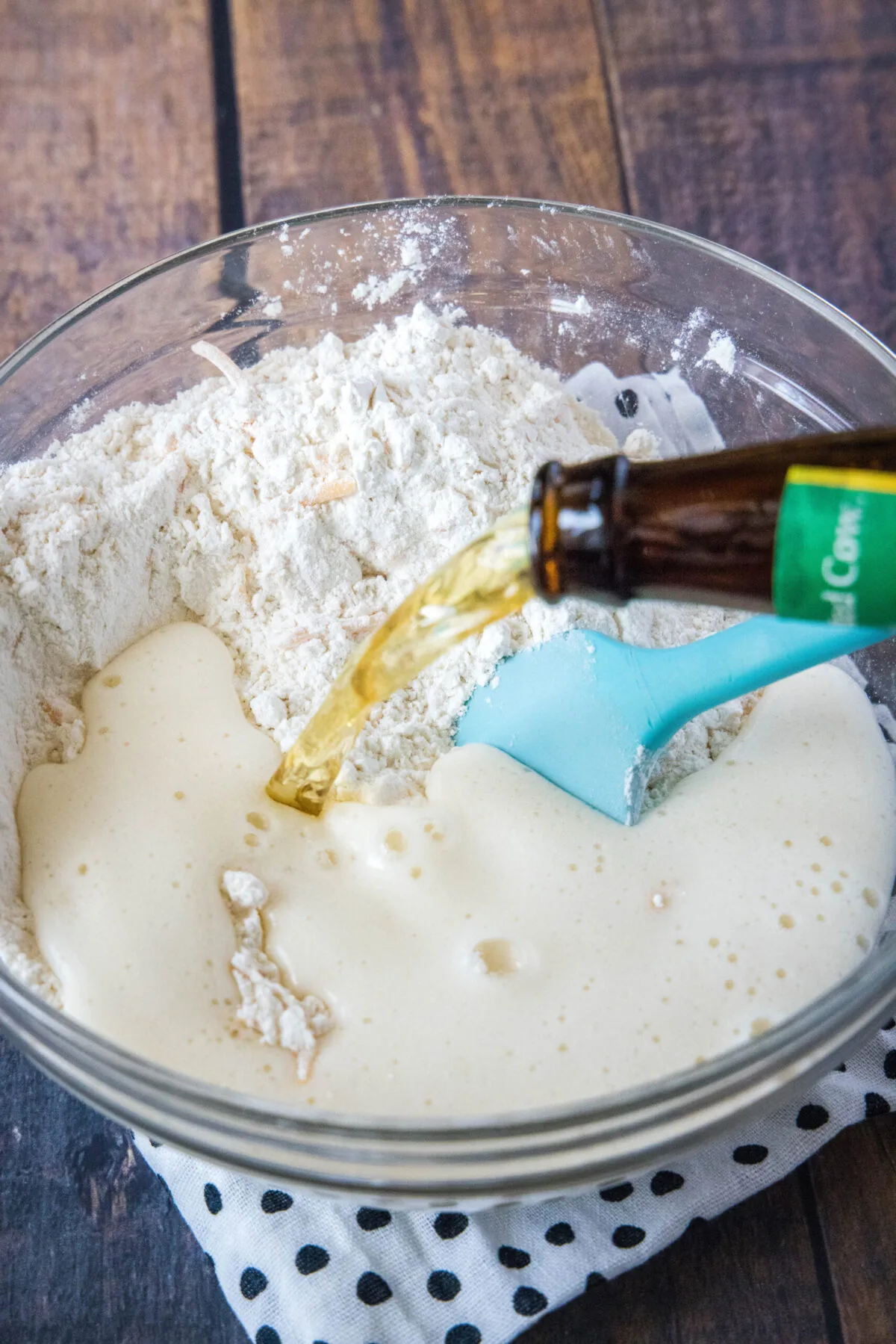 A bottle of beer being poured into a bowl of flour, cheese, and other ingredients, with a rubber spatula