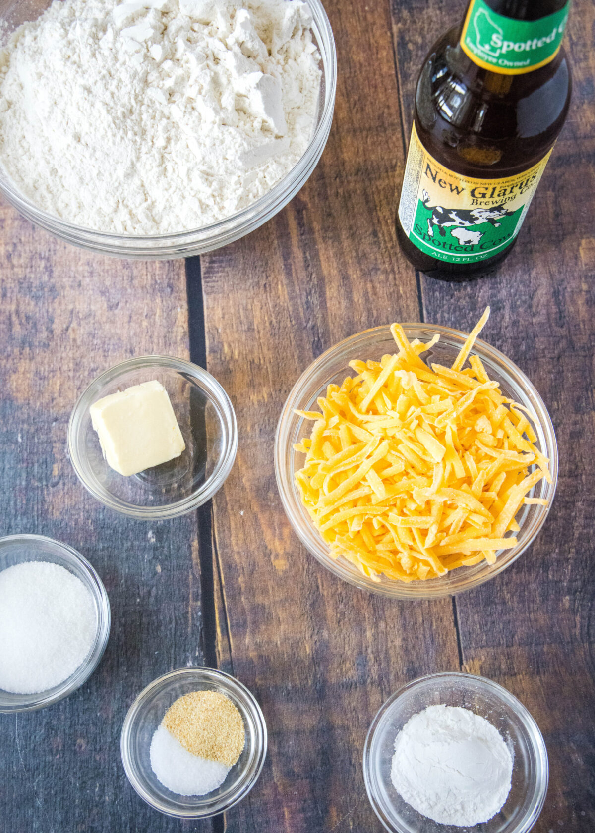 Overhead view of the ingredients needed for beer bread: a bowl of flour, a bowl of cheese, a bowl of baking powder, a bowl of sugar, a bowl of salt and garlic powder, a bowl of butter, and a bottle of beer