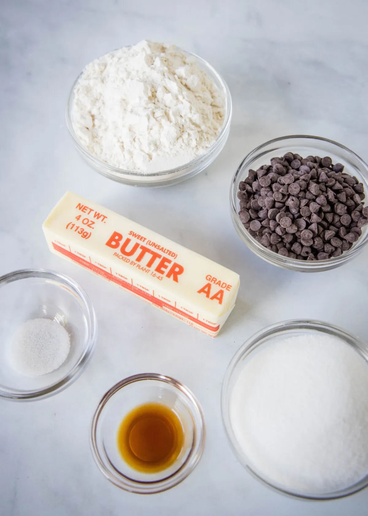 Overhead view of the ingredients needed for chocolate chip shortbread: a bowl of flour, a bowl of sugar, a bowl of chocolate chips, a bowl of salt, a bowl of vanilla, and a stick of butter.