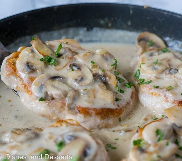 Close up of a spatula sliding under smothered pork chops with mushroom gravy in a skillet.