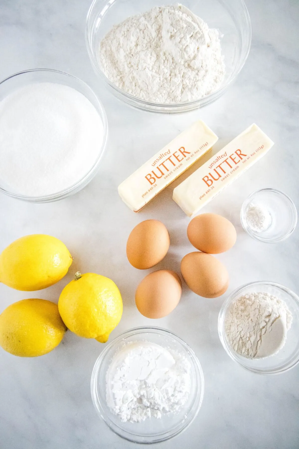 Overhead view of the ingredients needed for lemon bars: a big bowl of flour, a little bowl of flower, a bowl of sugar, a bowl of powdered sugar, a bowl of baking powder, two sticks of butter, four eggs, and three lemons
