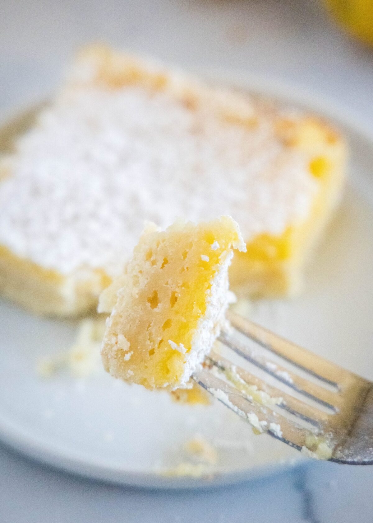 A bite of a lemon bar on a fork, with a lemon bar on a plate in the background